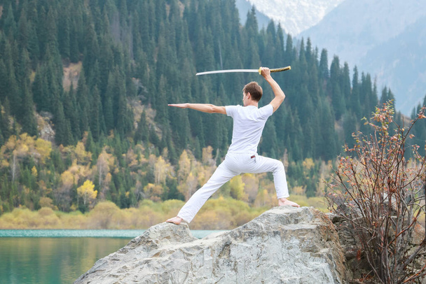 Ein junger Mann steht auf einem großen Stein und hält ein japanisches Schwert in seinen Händen. - Foto, Bild