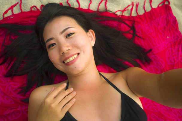 outdoors fresh and natural portrait of young beautiful and happy Asian Korean woman in bikini lying flat on sarong on beach sand taking selfie photo with mobile smiling cheerful and relaxed - Zdjęcie, obraz