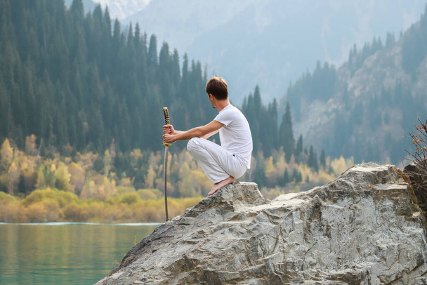 Ένας σοφός άνθρωπος κάθεται πάνω σε μια πέτρα και κρατά ένα σπαθί στα χέρια του. Ορεινή λίμνη φόντο - Φωτογραφία, εικόνα