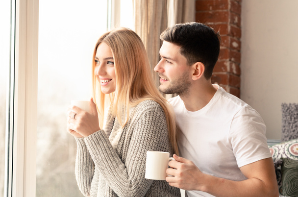Настоящая любовь. Улыбающиеся мужчина и женщина чашки кофе глядя в окно утром
 - Фото, изображение