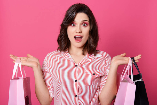 Portrait of young astonished lady standing with shopping bags in hands and amazedly looking directly at camera while posing isolated over pink background, dresses striped blouse, having dark hair. - Фото, изображение