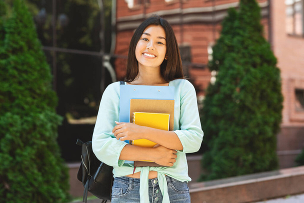 Smiling asian student girl with backpack and workbooks over college background - 写真・画像