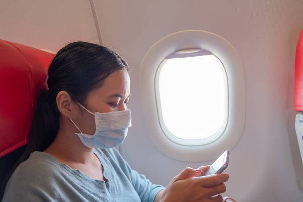 Chica asiática utilizar una máscara de protección para coronavirus o covid 19 en el aeropuerto. Tailandesa jugando con su teléfono en el avión
. - Foto, imagen