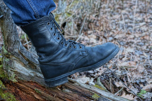ένα μαύρο ψηλό στρατιωτικό δερμάτινη μπότα σε ένα πόδι στέκεται σε ένα καφέ κούτσουρο δέντρο στη φύση - Φωτογραφία, εικόνα