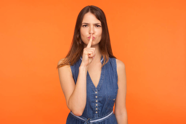 Be quiet, don't speak! Strict serious brunette woman in denim dress making gesture to keep silence, holding finger on lips, shushing hiding secret. indoor studio shot isolated on orange background - Foto, immagini