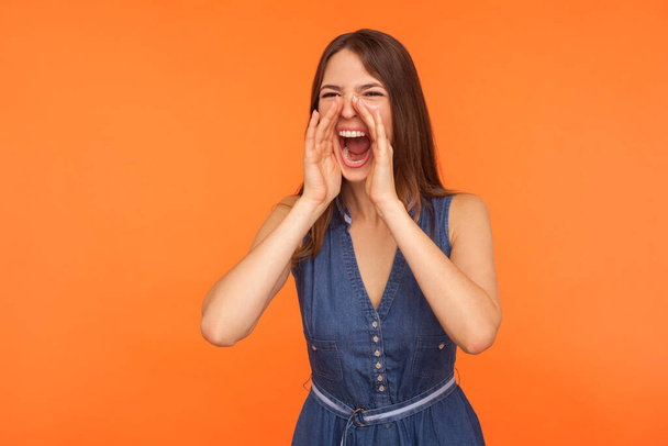 Brunette woman in denim dress keeping arms around wide open mouth, screaming loudly, calling attention to important news, announcing event, message. indoor studio shot isolated on orange background - Fotografie, Obrázek
