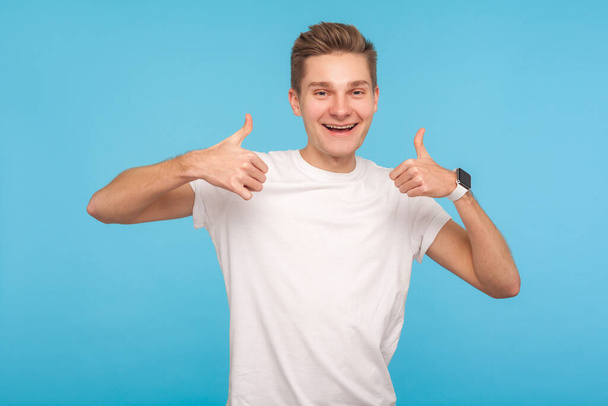 Gut gemacht! Porträt eines glücklichen, enthusiastischen Mannes im lässigen weißen T-Shirt, der die Daumen nach oben zeigt und freundlich lächelt und sich über das Ergebnis freut. Innenstudio isoliert auf blauem Hintergrund aufgenommen - Foto, Bild