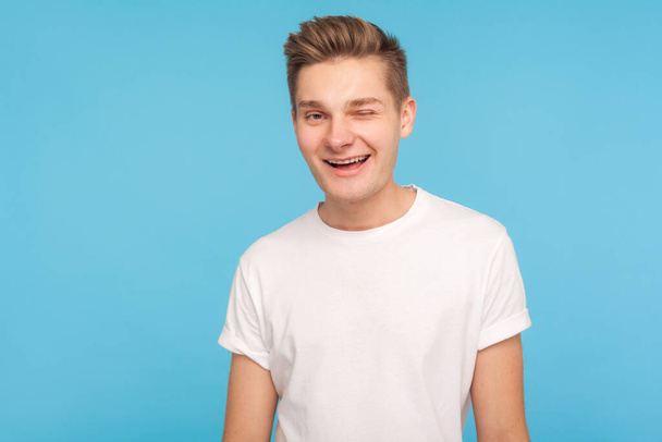 Portrait of carefree positive man in casual white t-shirt winking with playful funny expression, smiling flirting to camera, wants to invite on date. indoor studio shot isolated on blue background - Zdjęcie, obraz
