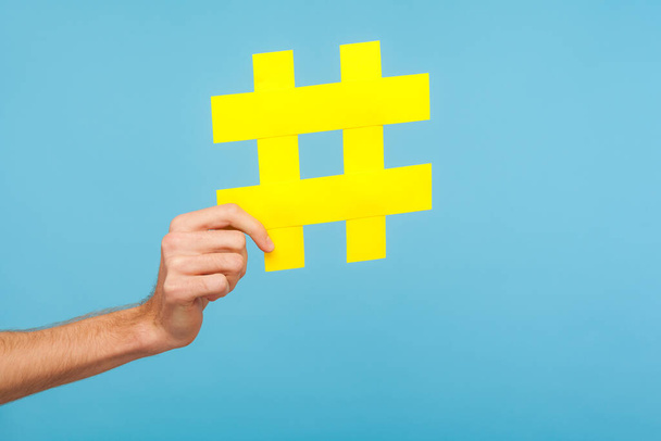 Closeup of male hand holding big yellow hash sign, hashtag symbol of internet trends and popular blogs, recommendation to follow social media content. indoor studio shot isolated on blue background - Photo, image