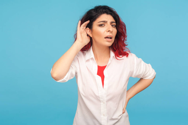 Portrait of nosy hipster woman with fancy red hair in shirt listening attentively holding hand near ear to hear confidential talk, hard to understand. indoor studio shot isolated on blue background - 写真・画像