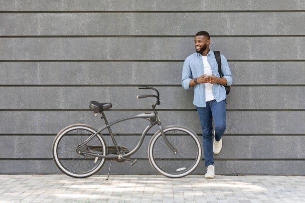 Akıllı telefonlu yakışıklı afro adam bisikletin yanında duruyor, duvara yaslanıyor. - Fotoğraf, Görsel