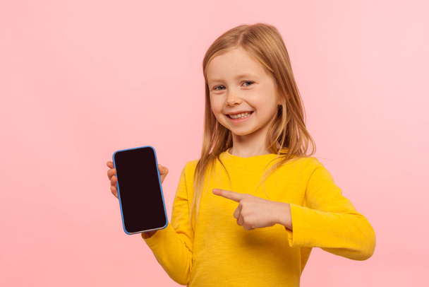 Look at my cellphone! Adorable glad little girl pointing smartphone and smiling at camera, advertising of mobile device, kid service, easy application. indoor studio shot isolated on pink background - Foto, Imagem