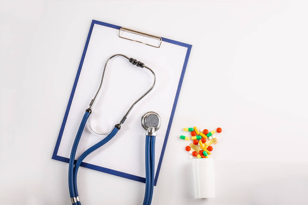Stethoscope, πρόχειρο και πολύχρωμα χάπια από το μπουκάλι σε λευκό φόντο απομονώνονται σε λευκό. Έννοια του ιού 2019-nCoV, coronavirus, ασθένεια. Ιατρικός έλεγχος. - Φωτογραφία, εικόνα
