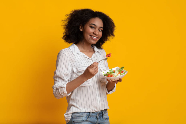 Mujer afro sonriente comiendo ensalada de verduras frescas de pie sobre fondo amarillo
 - Foto, imagen