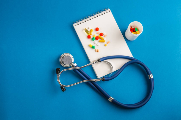 Stethoscope, σημειωματάριο και πολύχρωμα χάπια από το μπουκάλι σε μπλε φόντο. Έννοια του ιού 2019-nCoV, coronavirus, ασθένεια. Ιατρικός έλεγχος. - Φωτογραφία, εικόνα