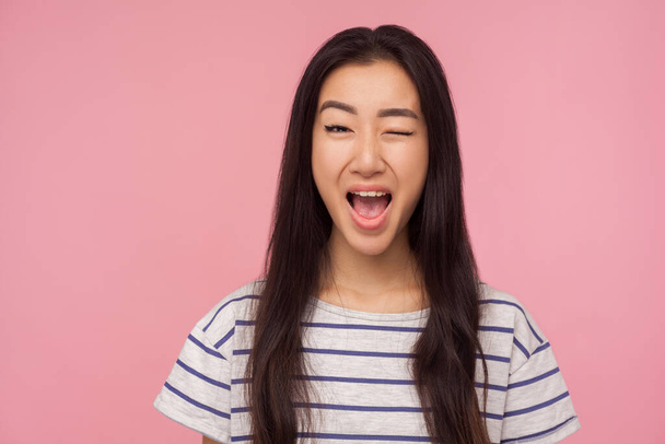 Portrait of playful girl with long hair winking at camera and smiling broadly, excited about acquaintance, having some cunning idea, blinking eye. indoor studio shot isolated on pink background - Foto, Imagem