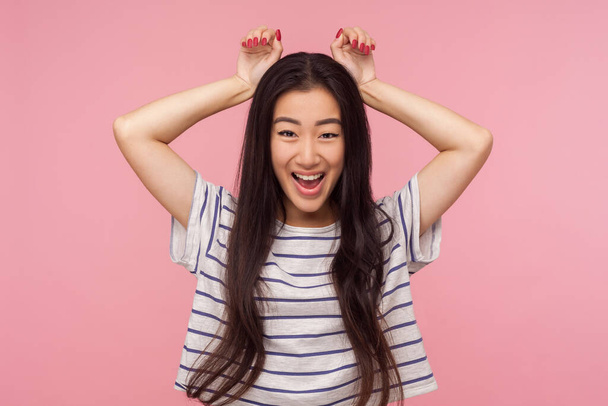 Portrait of childish funny girl with long brunette hair in striped t-shirt showing bunny ears gesture and smiling with playful carefree expression. indoor studio shot isolated on pink background - Foto, Imagen
