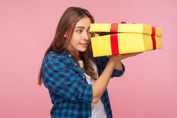 Portrait of curious cheerful girl in checkered shirt looking inside present box with nosy expression, peeking with interest, in anticipation of birthday surprise. indoor studio shot, pink background - Foto, Imagem