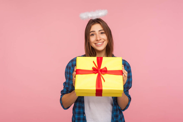 Present from angel. Portrait of generous happy beautiful girl with halo over head holding gift box and smiling kindly, congratulating on birthday. indoor studio shot isolated on pink background - Фото, изображение