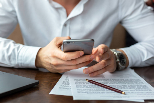 Closeup of hands of young businessman wearing a white fashion shirt with a mobile phone in a cafe with a laptop and documents. Freelance and selfemployment concept. Distance job - Photo, image