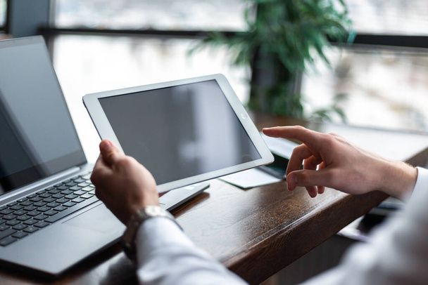 Close-up van de handen met een tablet met diagrammen van jonge zakenman dragen van een witte mode shirt in een cafe met een laptop en documenten. Kies en kies opties. Begrip freelance en zelfstandige - Foto, afbeelding