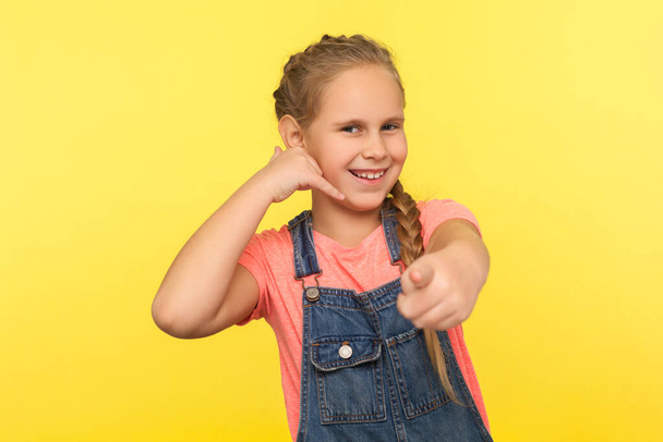 Hey you, let's communicate! Portrait of funny positive little girl with braid in denim overalls making call with fingers shaped like telephone and pointing to camera. indoor studio shot, isolated - Foto, Imagen
