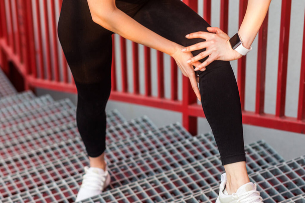 Closeup of female runner in sportswear holding painful knee while climbing stairs, touching leg suffering muscle strain, hurting to walk with sprained ligaments, joint injury at sports training. - Foto, afbeelding
