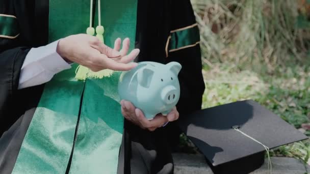 Die Person in der Bachelor-Uniform steckt Münzen in ein blaues Sparschwein, Sparen für Bildungsplan und Rentenfondskonzept. - Filmmaterial, Video