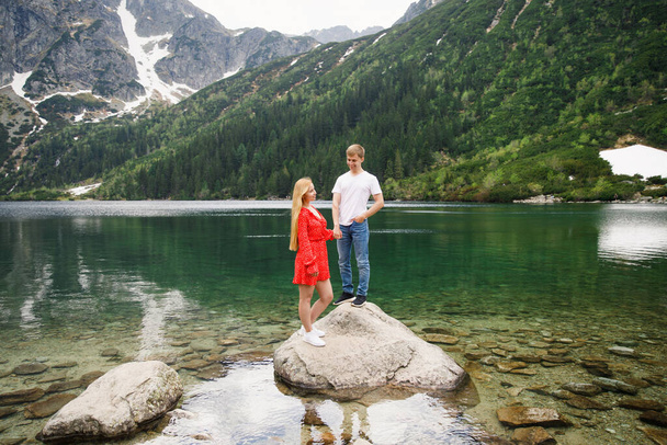 a young couple, a girl in a red dress, a man in a white T-shirt and blue pants stand on a background of mountains near the lake in the park - Fotografie, Obrázek