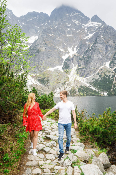 mladý pár, dívka v červených šatech, muž v bílém tričku a modrých kalhotách stojí na pozadí hor u jezera v parku - Fotografie, Obrázek