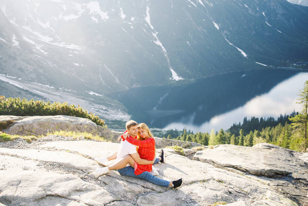 a young couple, a girl in a red dress, a man in a white T-shirt and blue pants stand on a background of mountains near the lake in the park - Foto, Imagem