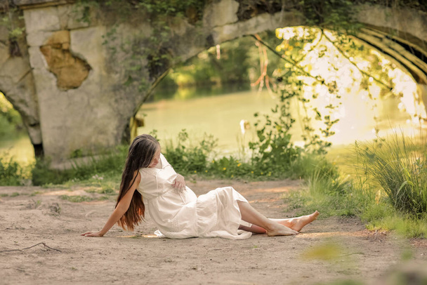 Ciężarna kobieta w długiej białej sukience spaceruje boso po lesie w pobliżu rzeki. - Zdjęcie, obraz