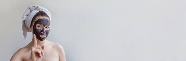 Reinigung der Haut Öko-Kosmetik Wellness-Entspannungskonzept. Schönheitsporträt einer jungen Frau mit Handtuch auf dem Kopf, die auf weißem Hintergrund eine schwarze Pflegemaske auf dem Gesicht trägt. Banner - Foto, Bild