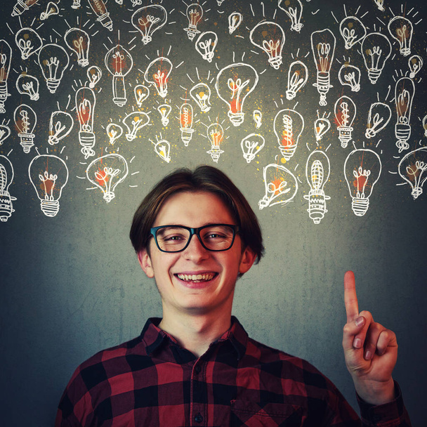 Joyful guy pointing index finger up, showing multiple light bulbs as creative ideas isolated on grey wall background. Positive thinking teenager, ingenious student find solutions to every problem. - Photo, Image