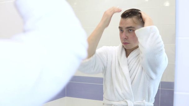 Un hombre joven aplica mousse para la colocación en el cabello. Un hombre con un albornoz blanco frente a un espejo de baño. Vista a través del espejo
 - Foto, Imagen