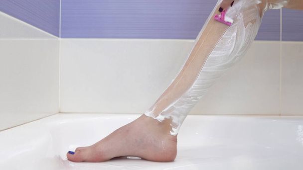 A young woman shaves hair on her legs. The girl applied shaving foam on her leg and started shaving. - 写真・画像