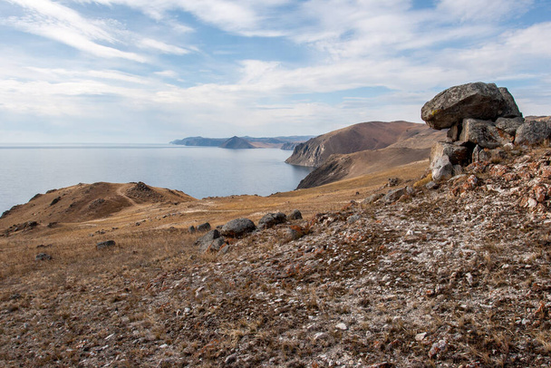 Brzeg jeziora Baikal z kamieniami na pierwszym planie. Wysoko skalisty brzeg. Na niebieskim niebie są chmury. Jezioro jest spokojne.. - Zdjęcie, obraz