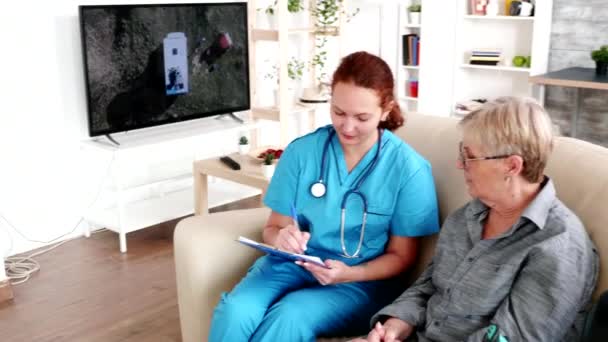 Nurse in retirement home taking notes on clipboard while talking with elderly retired woman - Video, Çekim