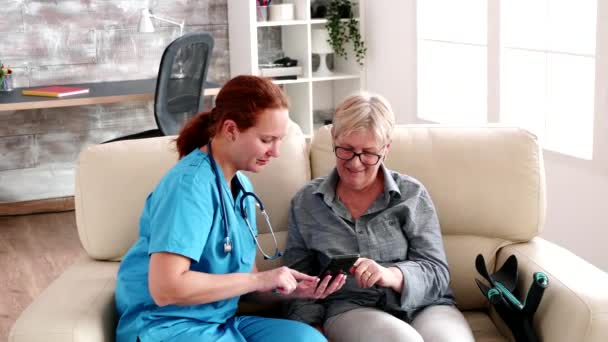 Huzurevindeki hemşire kanepede oturup yaşlı bir kadının akıllı telefon kullanmasına yardım ediyor. - Video, Çekim