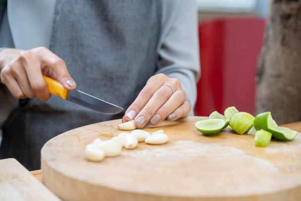 Asian female cut garlic by knife on the wood board with half lime beside it to preparing lunch at the kitchen. - Photo, Image
