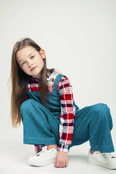 cute little girl sitting on the floor. Kid in a denim suit, in a plaid shirt and white sneakers. studio fashion photography. child fashion - Фото, зображення