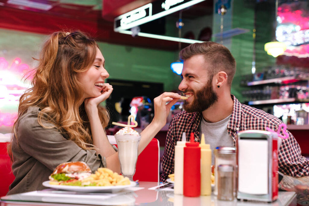 Image of a positive smiling young loving couple sit in retro bright cafe eat french fries indoors. - Fotografie, Obrázek