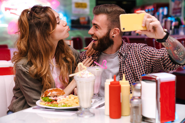Image of a happy cheery optimistic young loving couple sit in retro bright cafe take a selfie by mobile phone indoors. - Photo, image