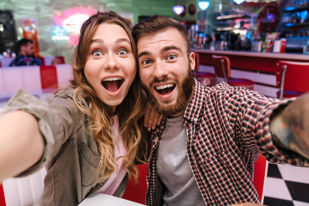 Image of a cute surprised excited young loving couple in retro bright street food cafe take a selfie by camera. - Foto, Imagem