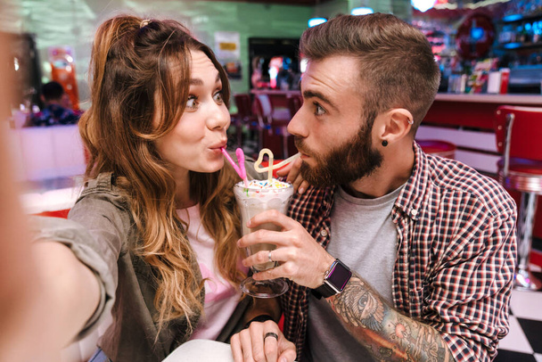 Imagen de una divertida pareja amorosa joven emocional positiva en retro café de comida de la calle brillante tomar una selfie por cámara bebiendo cóctel
. - Foto, imagen