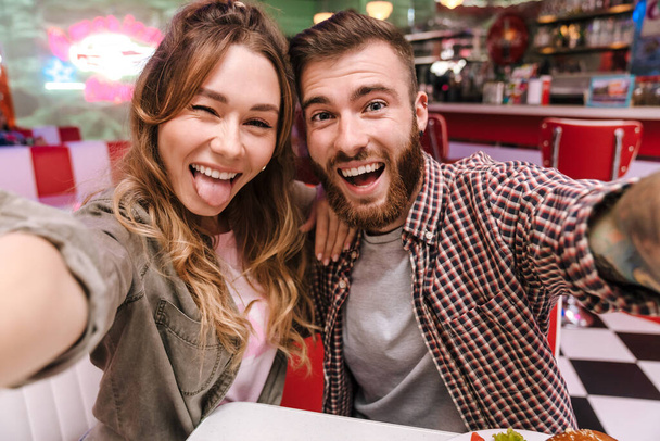 Image of a cute happy young loving couple in retro bright street food cafe take a selfie by camera showing tongue. - Foto, Bild