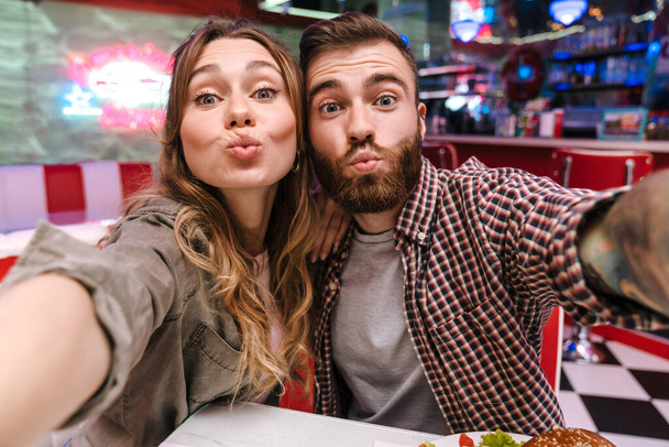 Image of a cute positive young loving couple in retro bright street food cafe take a selfie by camera blowing kisses. - Foto, immagini