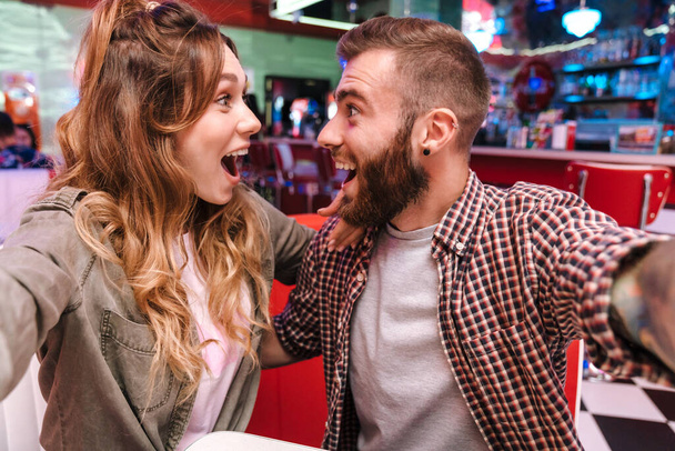 Image of a cute screaming happy young loving couple in retro bright street food cafe take a selfie by camera. - Zdjęcie, obraz