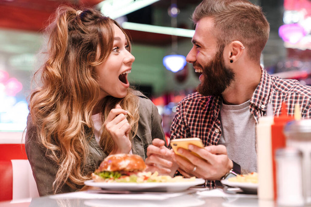Photo of a cute surprised screaming young loving couple in retro bright street food cafe using mobile phone. - Foto, immagini