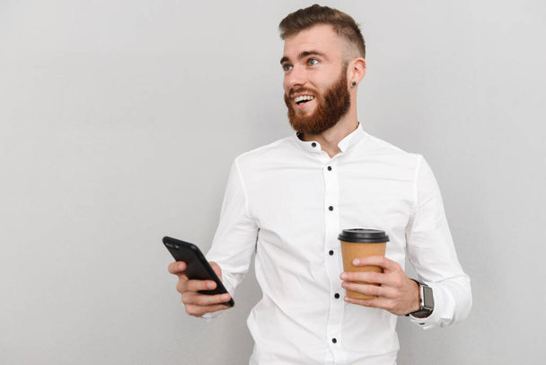 Portrait of an attractive handsome young businessman standing over gray background, using mobile phone while drinking takeaway coffee - Zdjęcie, obraz